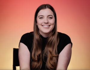 A headshot of Nicole Gress. She has long auburn hair and is wearing a black shirt. She is depicted against an orange and yellow ombre background.