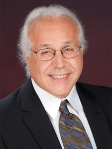 A headshot of Dr. Jeffrey Birnbaum who is wearing a black suit, white shirt, and patterned tie.