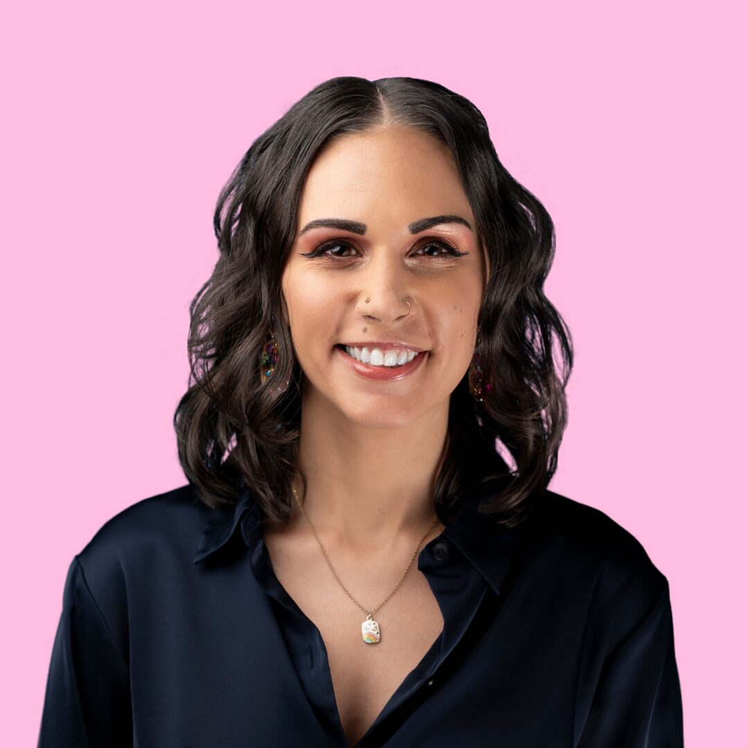 A headshot of Shohreh Davoodi wearing a navy blue satin collared shirt. Her brunette hair is shoulder length and wavy, and she is smiling against a pink backdrop.