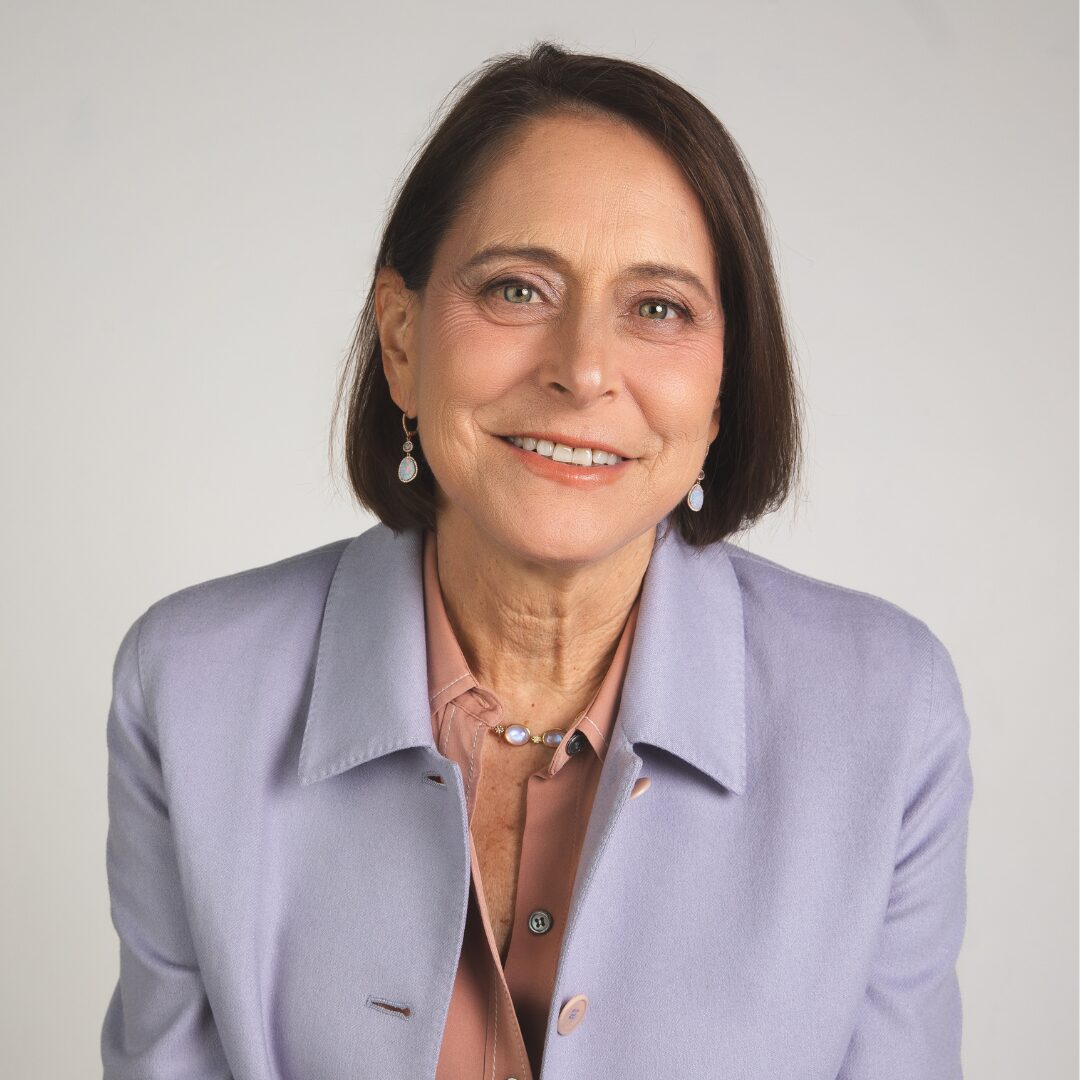 A headshot of Anne Miller, the PFLAG NYC Board Chair. She wears a coral colored shirt with a lavender blazer over it and sparkly hanging opal earrings.