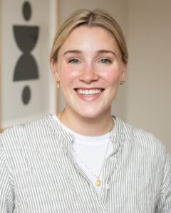 A headshot of Beth Gladstone, a woman with blonde hair and blue eyes wearing a tan and white striped blouse over a white t-shirt.