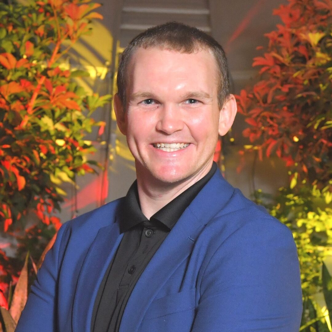A headshot of Brett P. Harrison, a PFLAG NYC Board Member. He wears a blue suit jacket over a black button up shirt.