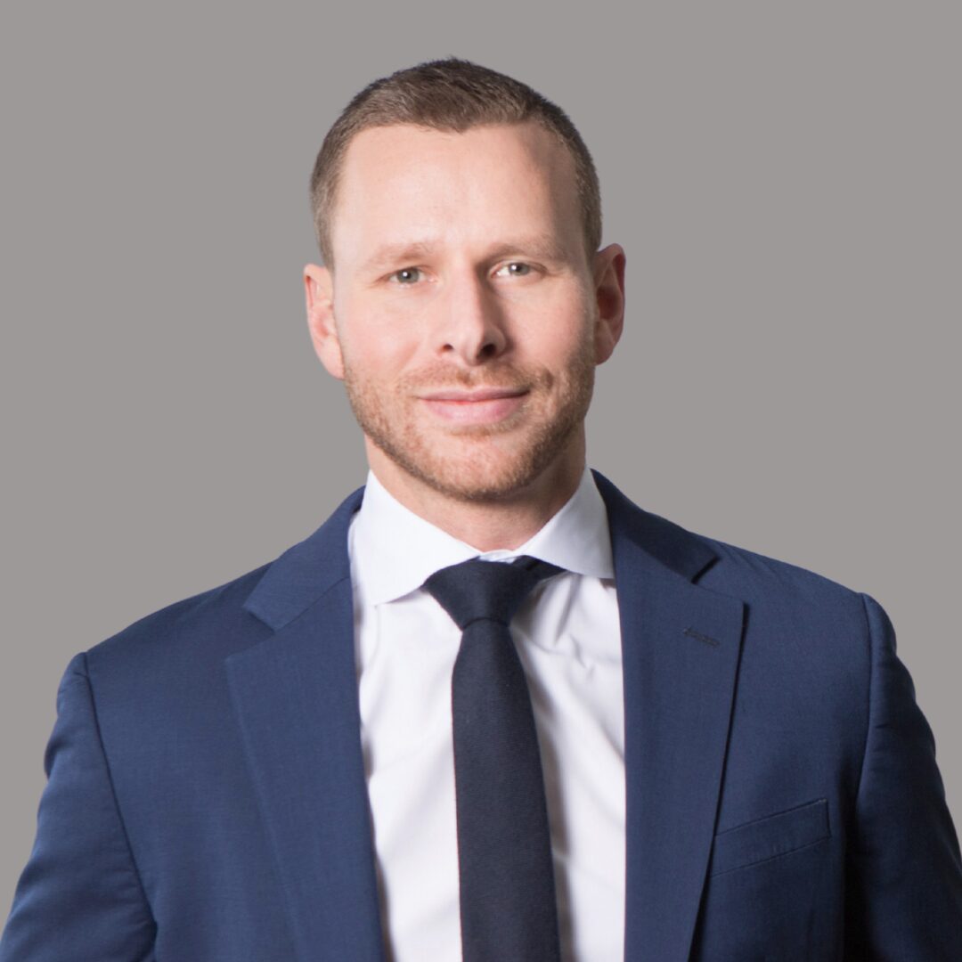 A headshot of PFLAG NYC Board Member Ralph Bassett. He wears a blue suit, white shirt, and navy tie.