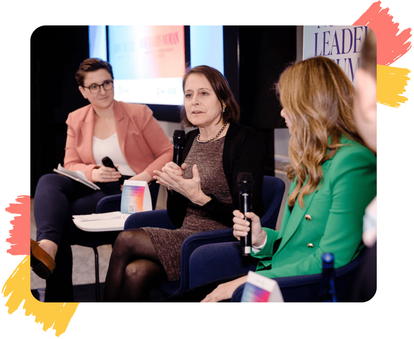 Four people seated in a panel discussion at an event. PFLAG NYC's Board Chair, Anne, is in a maroon dress speaking while holding a microphone. Another person in a green blazer listens attentively, and two others, including one with short hair and glasses in a salmon blazer, are engaged in the conversation. Event signage is partially visible in the background.