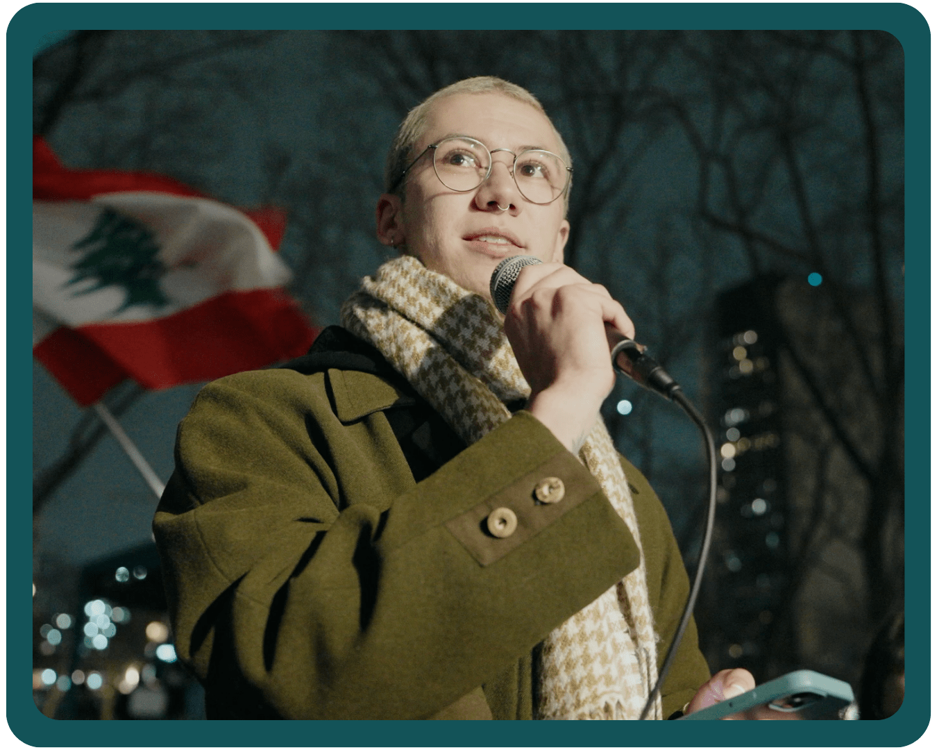 A photo of PFLAG NYC's Executive Director, Clark, speaking into a microphone outside at a rally while wearing an olive-colored coat and oatmeal-colored scarf.