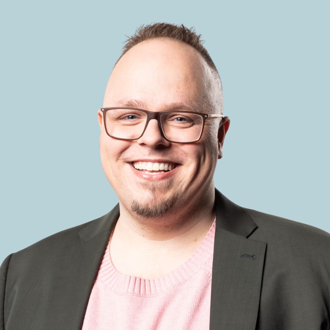 A headshot of PFLAG NYC Board Member Dr. Jared Fox wearing a gray suit jacket and pale pink sweater against a blue backdrop.
