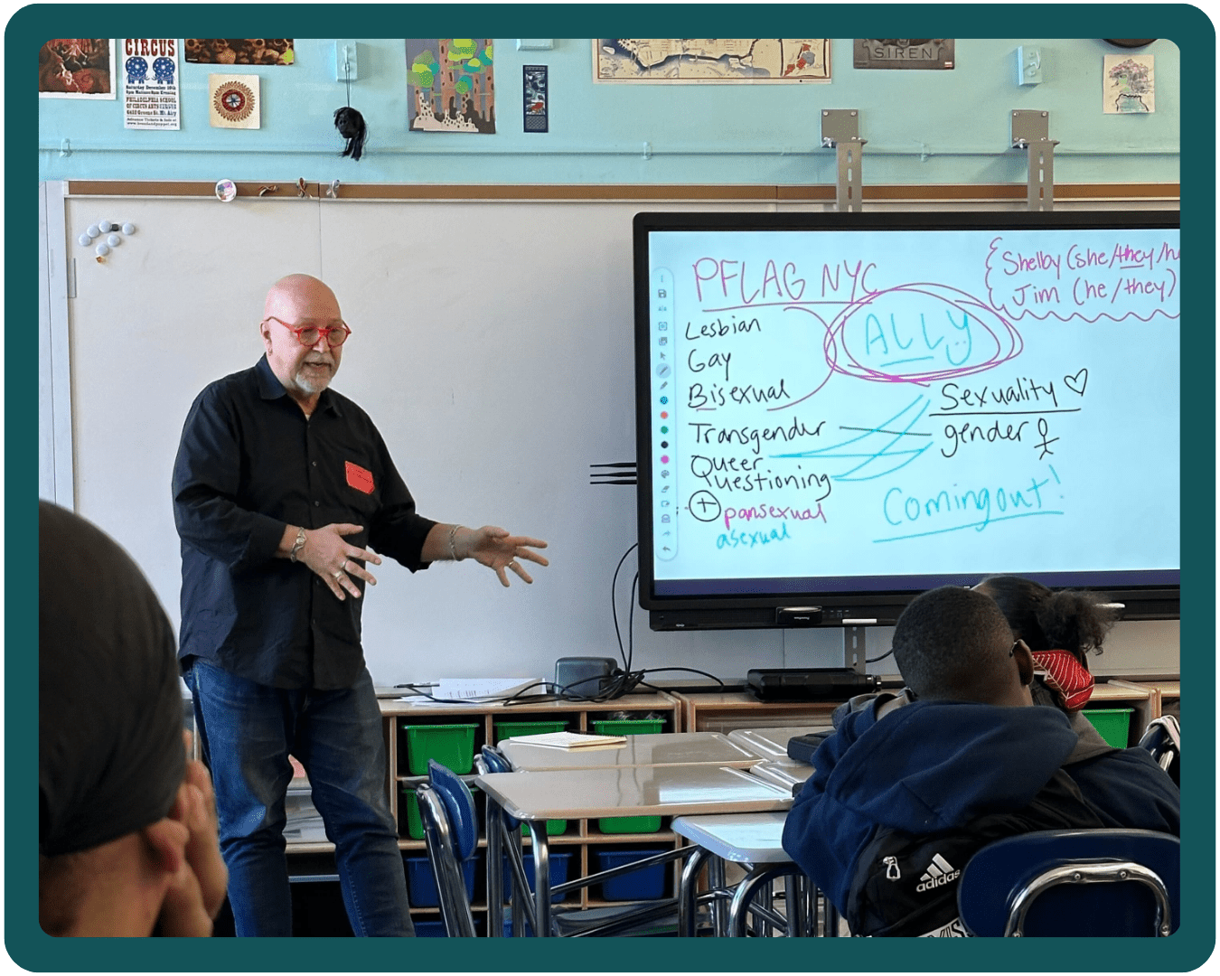 A Safe Schools Program volunteer stands at the front of a classroom and gestures to an electronic whiteboard that has words on it, including PFLAG NYC, ally, sexuality, gender, and coming out.