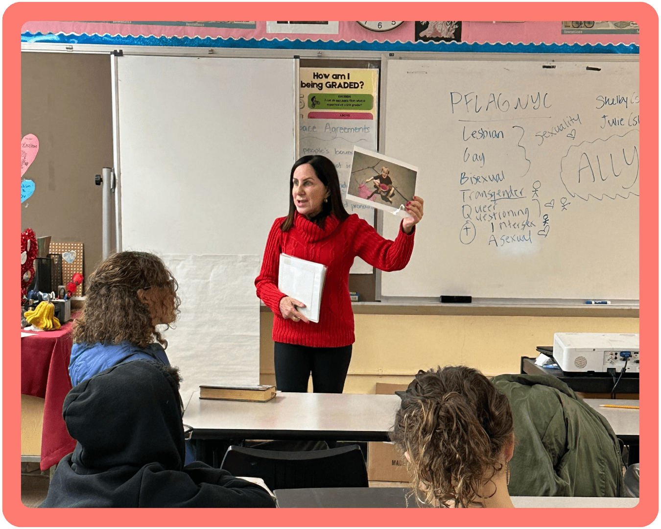 A Safe Schools Program volunteer wearing a bright red sweater stands at the front of a classroom holding up a photo of her child. Behind her is a whiteboard that has words written on it, including PFLAG NYC, ally, sexuality, and gender.