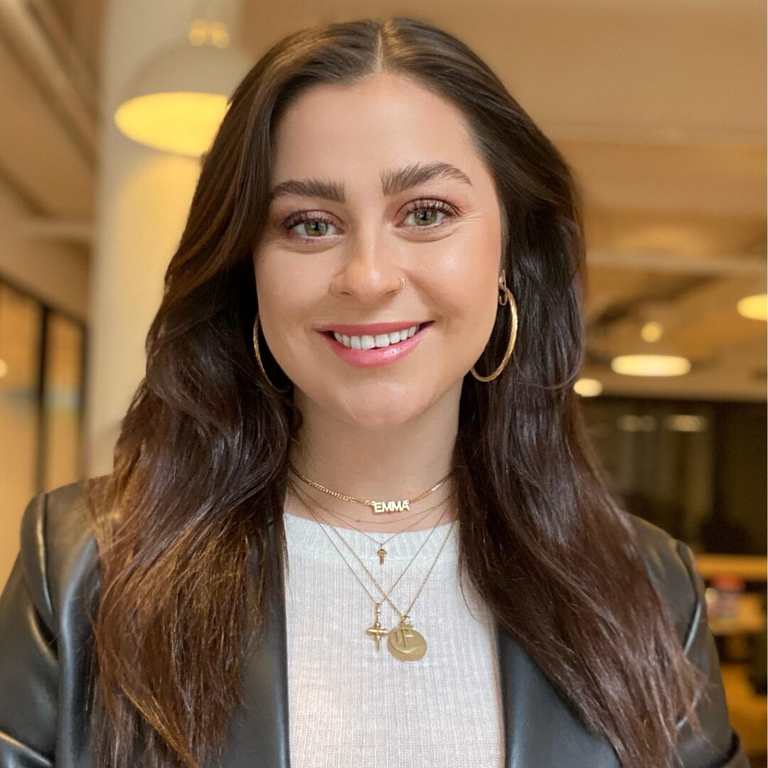 A headshot of Board Member Emma Ashworth. She smiles for the camera while wearing a white shirt and gold necklaces under a black leather jacket.