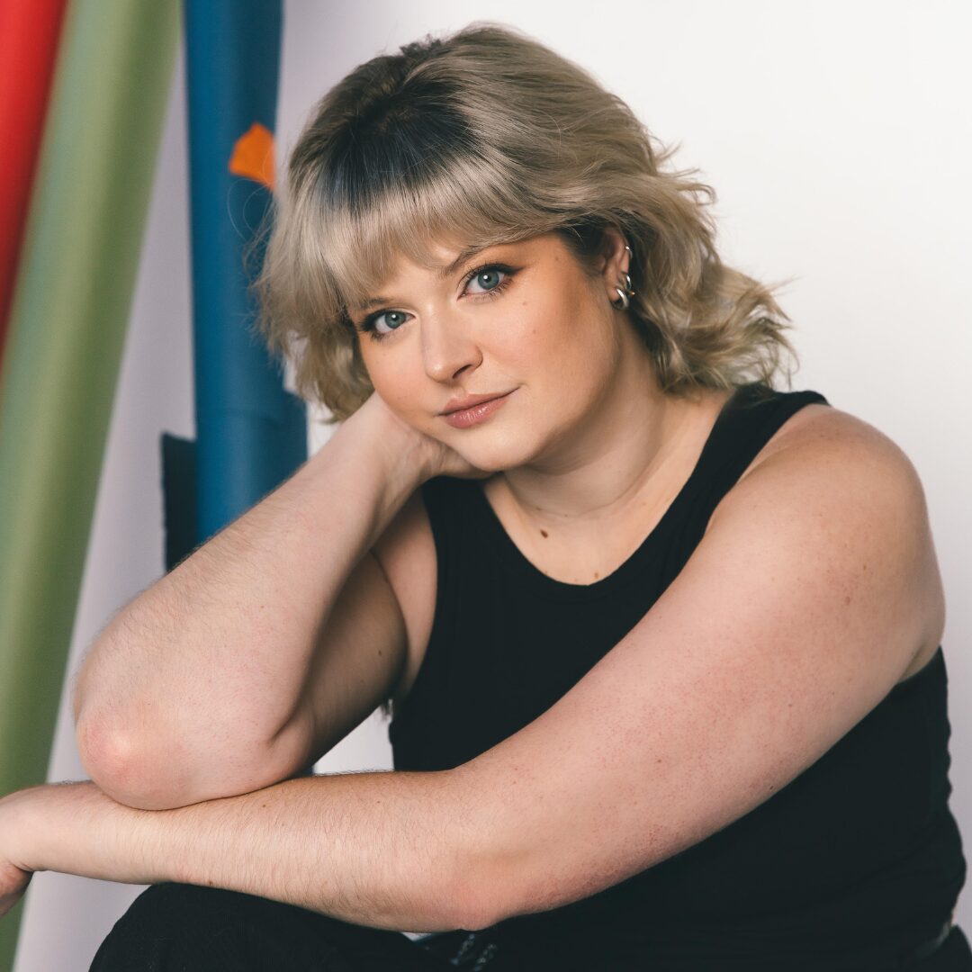 A headshot of Board Member McKenna C. Poe shows them in an all black ensemble with one arm across their knees and their other arm propping up their head.