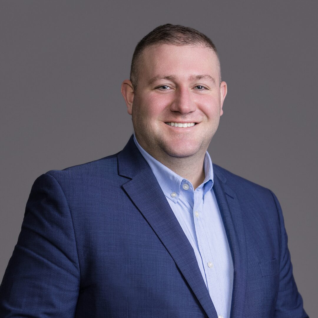 A headshot of Board Member Pat Bunting smiling and wearing a light blue collared shirt with a dark blue suit jacket over it.
