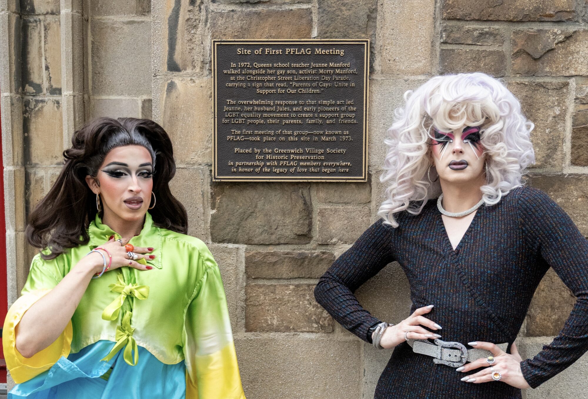 Drag queens Nancy NoGood and Reina NoBuena stand against a brick wall on either side of a plaque describing the location as the site of the first PFLAG meeting.