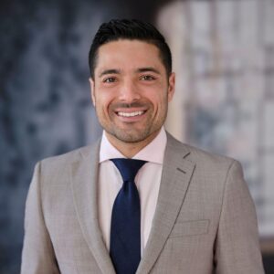 A headshot of Dr. Drew Marano who is smiling and wearing a tan suit jacket, collared shirt, and navy tie.