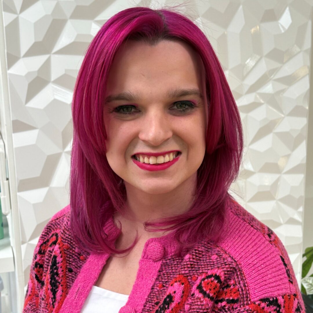 A headshot of board member Maxine Cline smiling. She has magenta hair and wears pink lipstick and a pink jacket over a white blouse.